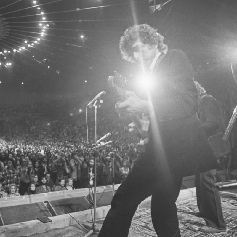 Young Bob Dylan performing on stage with a bright camera flash.